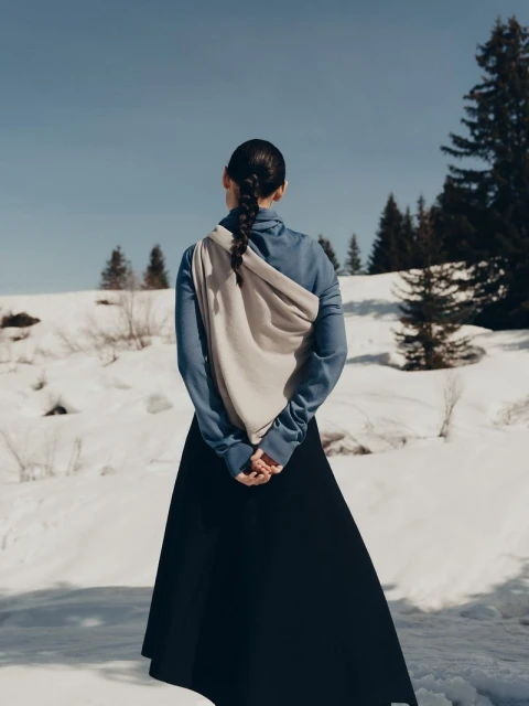 Woman in a black dress and blue top stands in a snowy field with evergreen trees under a blue sky in a fashion shot, with back to camera.