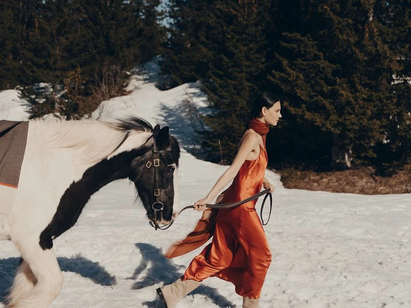 Woman in orange dress and beige boots walking alongside a black-and-white horse in snowy field, backed by evergreen trees and bright light.