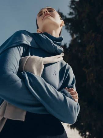 Model wearing a blue turtleneck sweater with a light-colored sweater draped over shoulders, captured against a clear sky