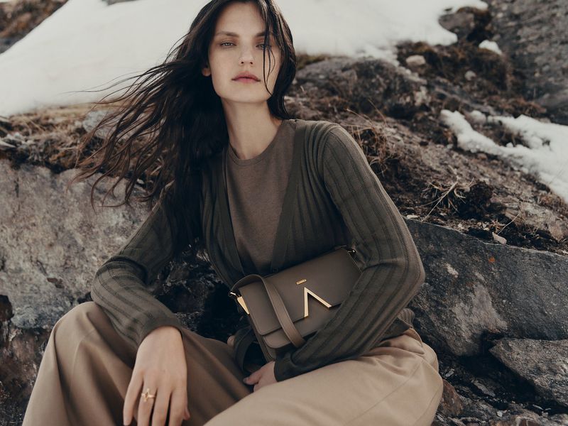 Model wearing olive cardigan and khaki trousers holding a leather shoulder bag outdoors against rocks with snow patches.