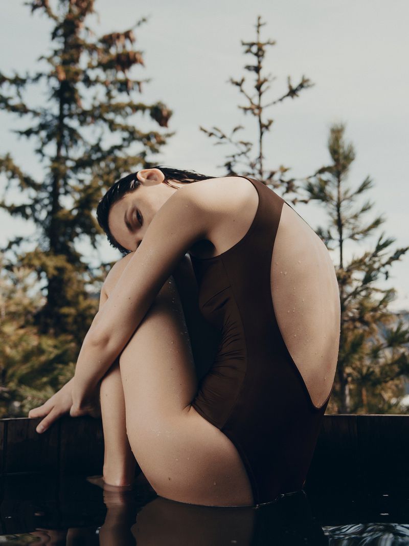 Woman in brown one-piece bathing suit sitting in water outdoors with evergreen trees in background. Natural lighting, medium shot.