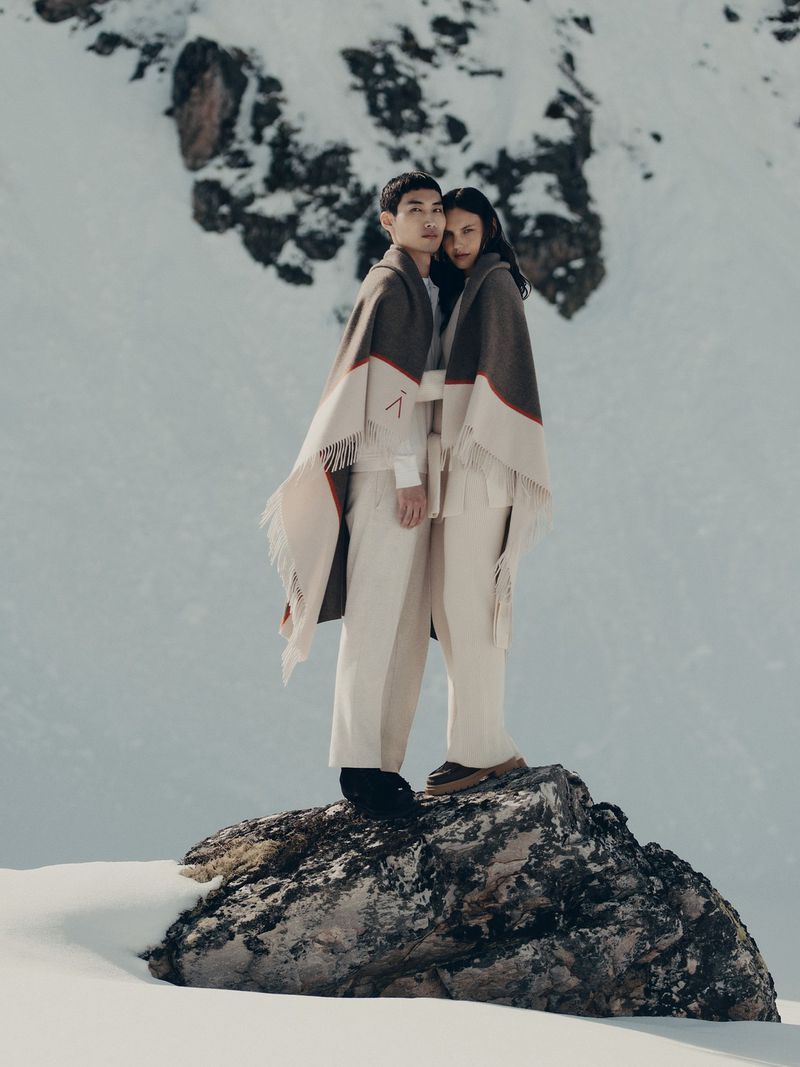 Two people, an Asian man and a woman, in matching capes and pants stand on a rock in a snowy mountain landscape, photographed in a medium shot.
