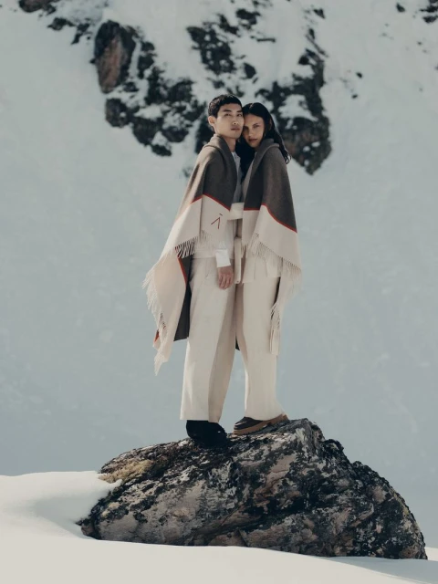 Two people, an Asian man and a woman, in matching capes and pants stand on a rock in a snowy mountain landscape, photographed in a medium shot.