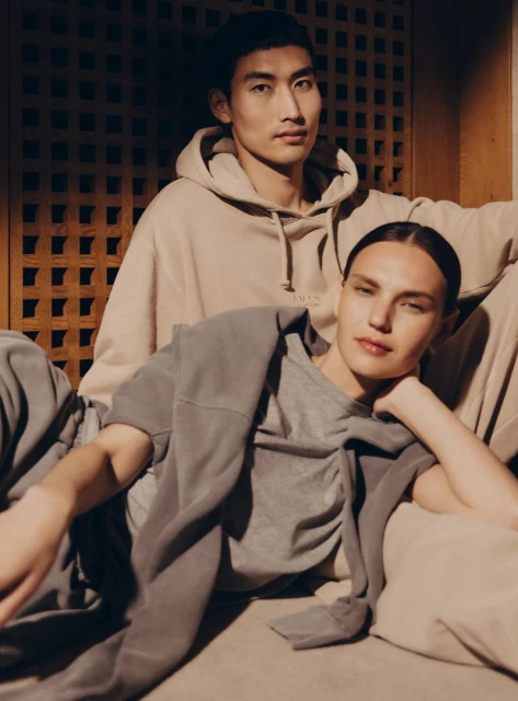 Two fashion models in casual clothes pose indoors, with a beige hoodie and grey sweatshirt. A soft, neutral color palette dominates.