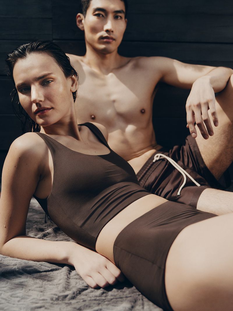 Two models reclining together outdoors, a woman in a brown set in the foreground and a shirtless man behind her