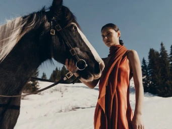 Model wearing a rust-orange dress posing next to a dark horse in a snowy landscape captured by Charlotte Lapalus.