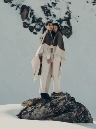 Two models wrapped in wool blankets standing on a snowy rock outcrop for Aman Essentials Heritage Collection Spring 2025.