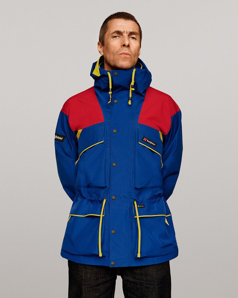 Liam Gallagher posing in a blue, red, and yellow color-blocked Berghaus jacket against a neutral grey studio background