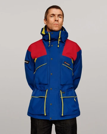 Liam Gallagher posing in a blue, red, and yellow color-blocked Berghaus jacket against a neutral grey studio background