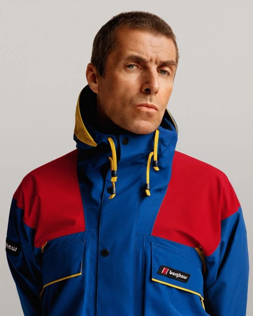 Portrait of a man in a vibrant blue, red, and yellow hooded jacket with Berghaus logo against a plain studio backdrop.