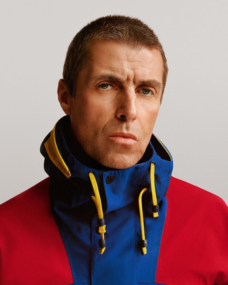 Close-up portrait of an adult male with short hair, wearing a red, blue, and yellow hooded jacket, set against a solid light background.