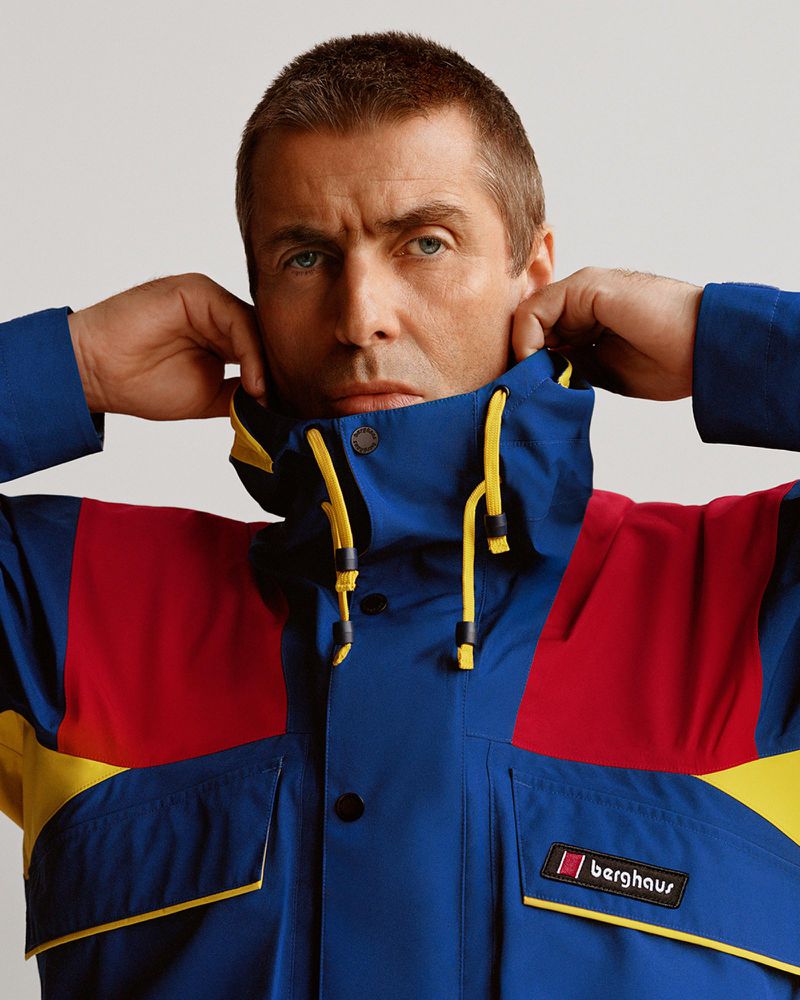 Close-up portrait of an adult man in a blue, red, and yellow windbreaker with a hood, looking directly at the camera with a neutral expression.