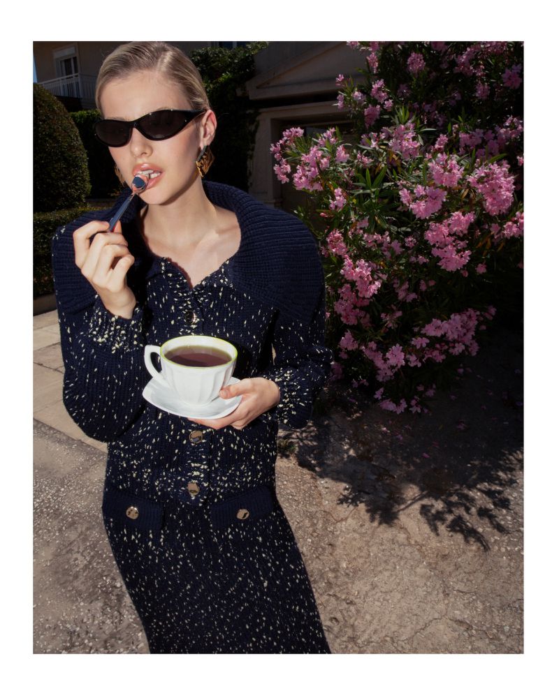 Elegant blonde woman in sunglasses sips tea outdoors. She wears a navy knit dress and holds a teacup and saucer. Pink flowers bloom nearby.