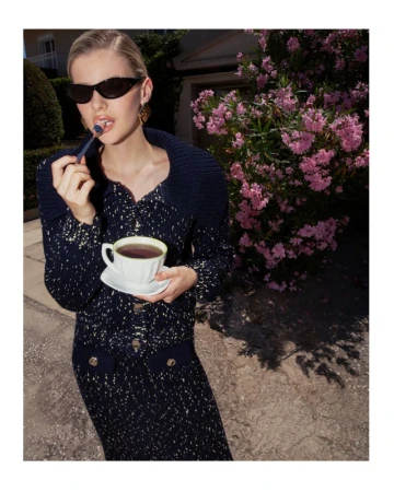 Elegant blonde woman in sunglasses sips tea outdoors. She wears a navy knit dress and holds a teacup and saucer. Pink flowers bloom nearby.