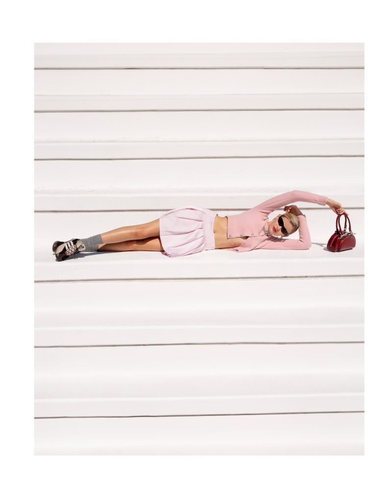 A young adult woman with sunglasses lays horizontally on white stairs in a pink skirt and matching cropped top, holding a red handbag.