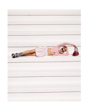 A young adult woman with sunglasses lays horizontally on white stairs in a pink skirt and matching cropped top, holding a red handbag.