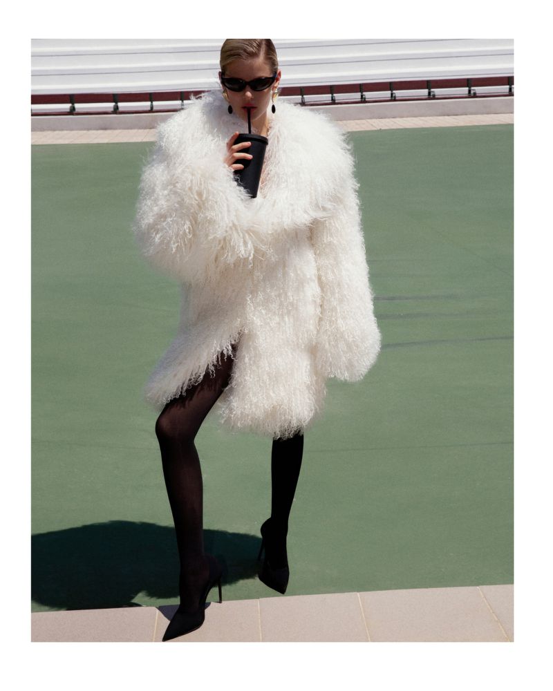 A blonde woman in a white coat and sunglasses drinks from a black cup while posing on a tennis court in a fashion editorial.