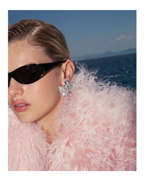 Close-up of a blonde adult female wearing sunglasses and large crystal earrings, posing in front of the ocean.