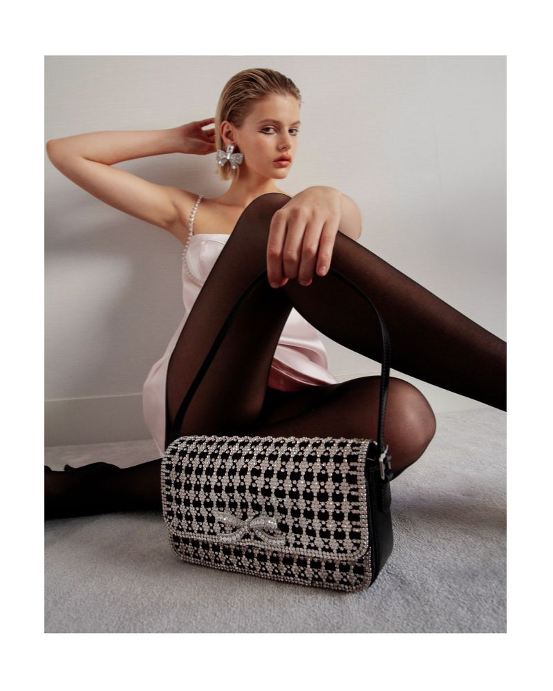 Model in a pink slip dress and black tights holding a crystal-embellished black handbag against a neutral wall.