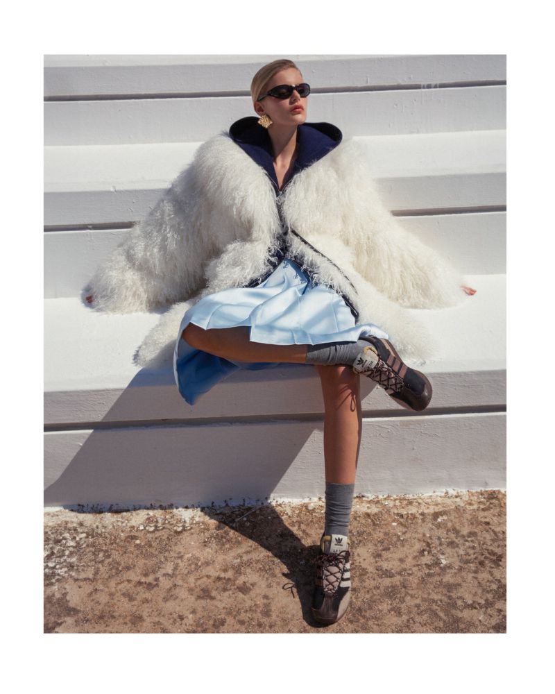 Stylish woman in white fur coat, blue dress, sneakers and sunglasses poses on white stairs outside on a sunny day in a fashion shot.
