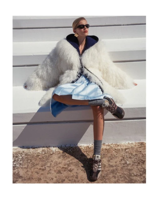 Stylish woman in white fur coat, blue dress, sneakers and sunglasses poses on white stairs outside on a sunny day in a fashion shot.