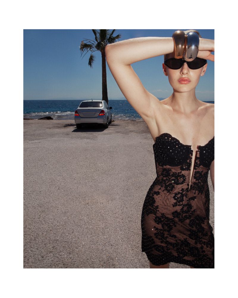 Model in black lace dress with metallic cuffs and sunglasses posing by the coast with a parked car and palm tree.