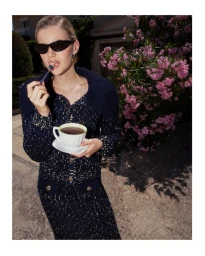 Model wearing dark sunglasses and a dark tweed knit cardigan holding a teacup outdoors beside a bush of pink flowers.