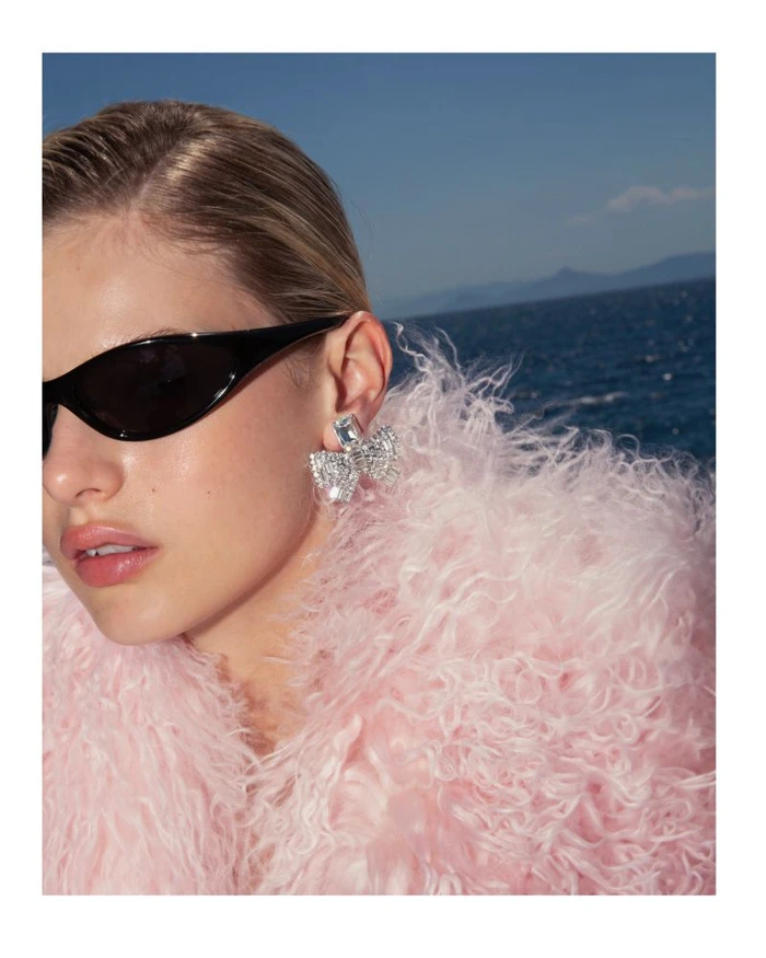 Close-up portrait of a woman wearing black sunglasses, diamond bow earrings, and a textured pink fuzzy coat against an ocean backdrop.