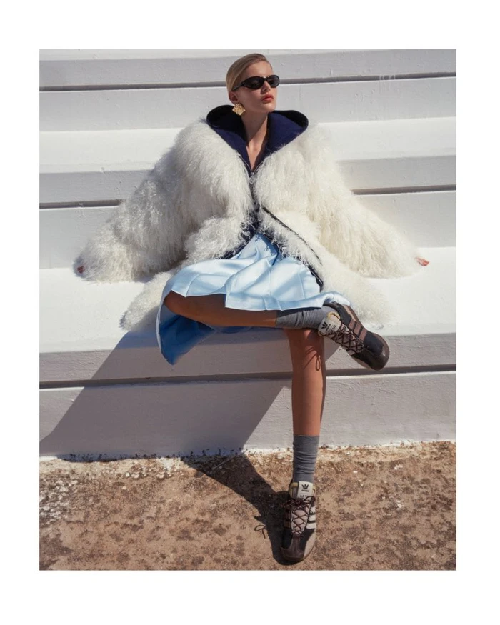Model in white shaggy coat and blue skirt seated on white outdoor tiered seating wearing sunglasses