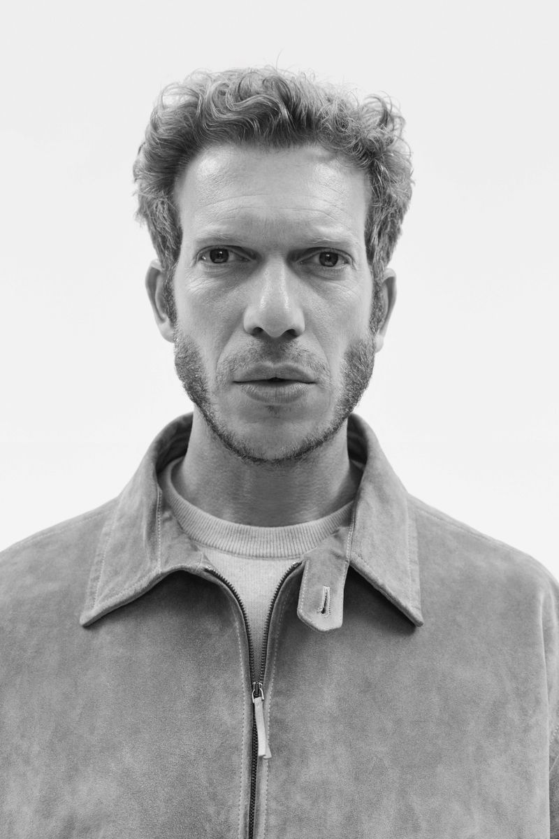 Black-and-white portrait of an adult male with curly hair and sideburns wearing a jacket, looking directly at the camera in a studio.