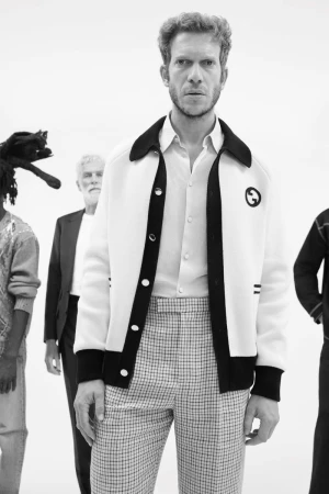 Monochrome fashion portrait of a male model wearing a button-up shirt, patterned trousers, and a contrasting jacket against a plain white studio background.
