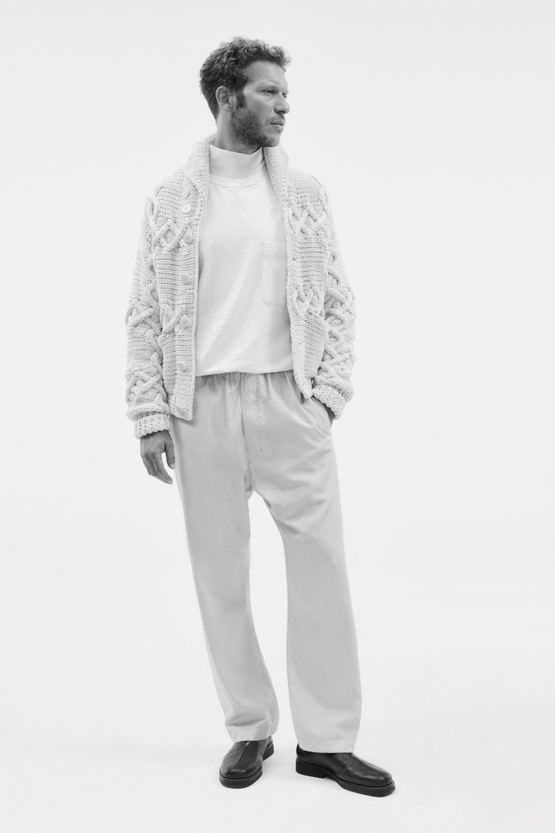 Full-length studio shot of a man in a white shirt, cable-knit cardigan, and light-colored pants, looking to the side with a neutral expression.