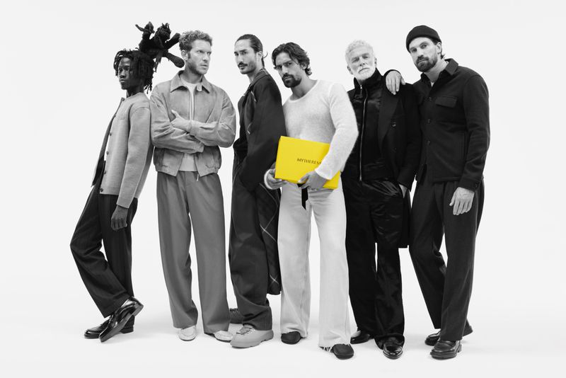 Group of six men in casual fashion, posed together in a bright studio, one holding a yellow MYSTAYS branded box, black and white.
