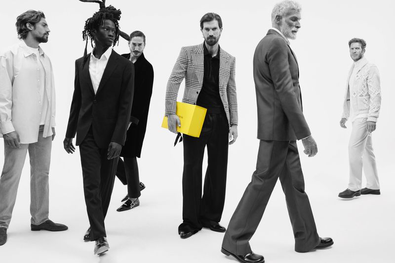 Editorial fashion shot: group of diverse men in suits walking on a white background, one carrying a yellow box, minimalist style.