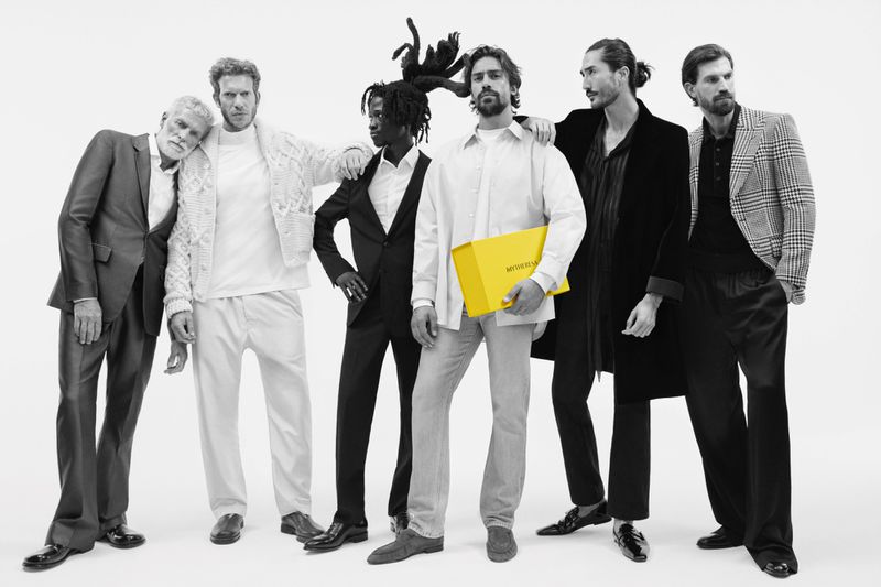 Group of six stylish men in an indoor photo studio; one man holds a yellow box that reads, 'MYSTERYLAND.' Black and white image with one color pop.