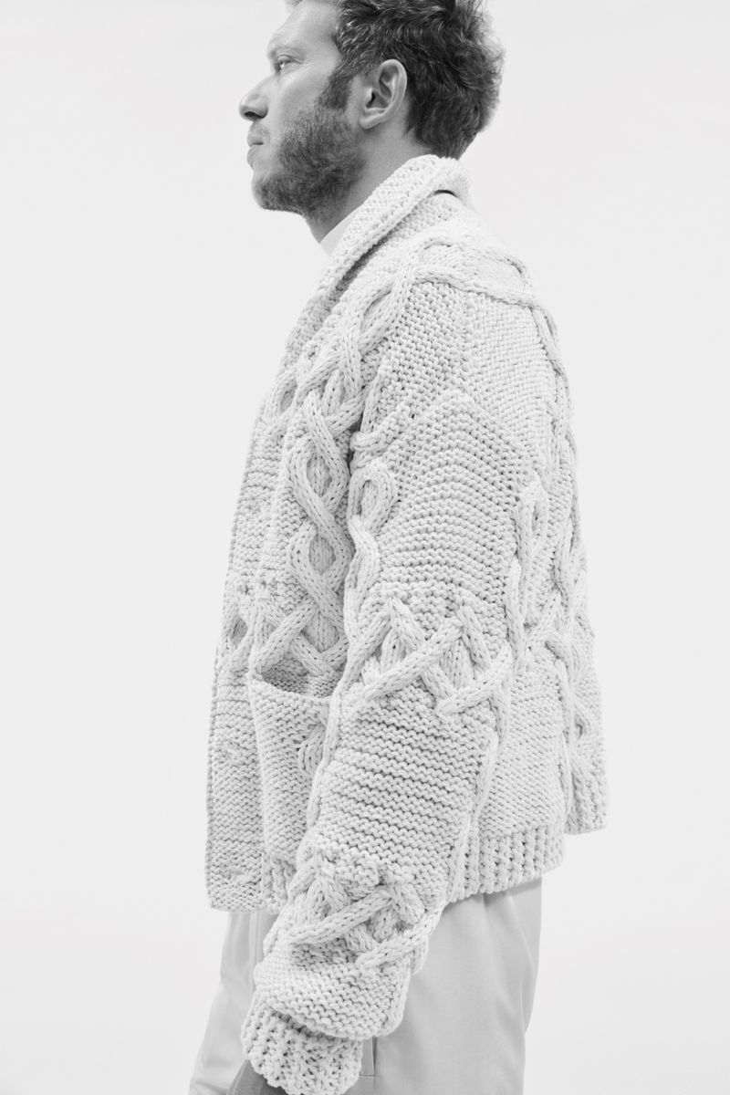 Monochrome portrait of a man in a cable knit cardigan against a light backdrop, showcasing fashion and texture. Profile view.