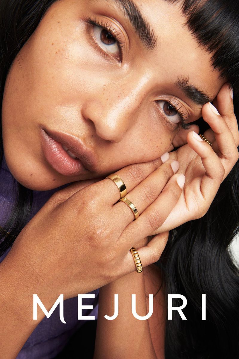 Close-up of a young adult female model with gold rings on her fingers, wearing a purple top and black hair, with the MEJURI logo.