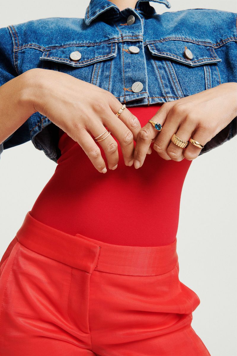 Close-up of hands wearing gold rings, styled with a cropped denim jacket, red top, and red trousers on white background.