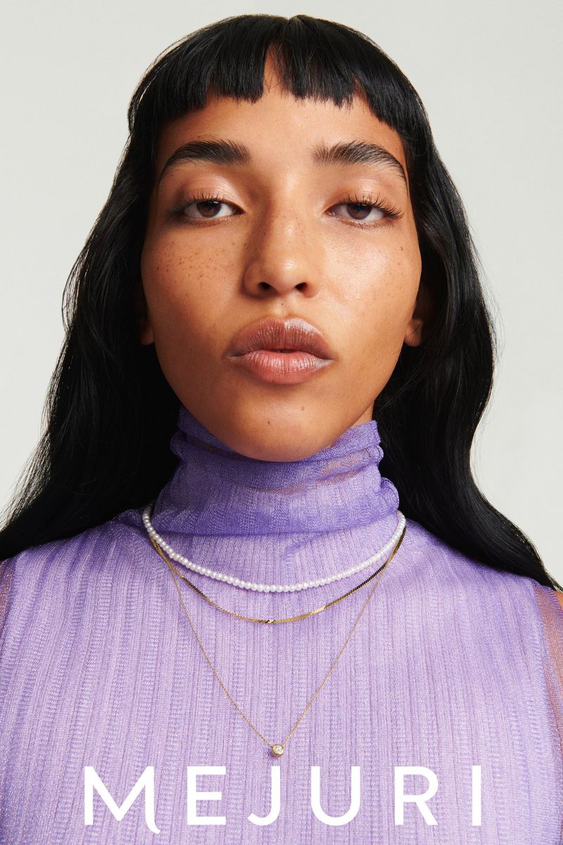 Close-up of an adult female wearing layered gold and pearl necklaces, styled with a sheer lilac top and dark hair against a plain backdrop.