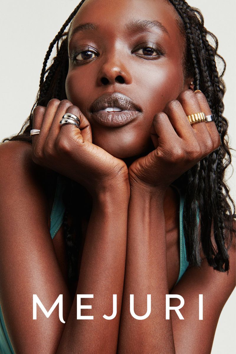 Close-up portrait of a Black woman resting her chin on her hands, showcasing stacked silver and gold rings against a neutral background.