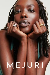 Close-up portrait of a Black woman resting her chin on her hands, showcasing stacked silver and gold rings against a neutral background.