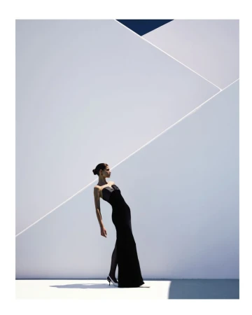 Elegant woman in a black strapless gown gracefully poses against a geometric white wall, accentuated by shadows and a touch of blue sky.