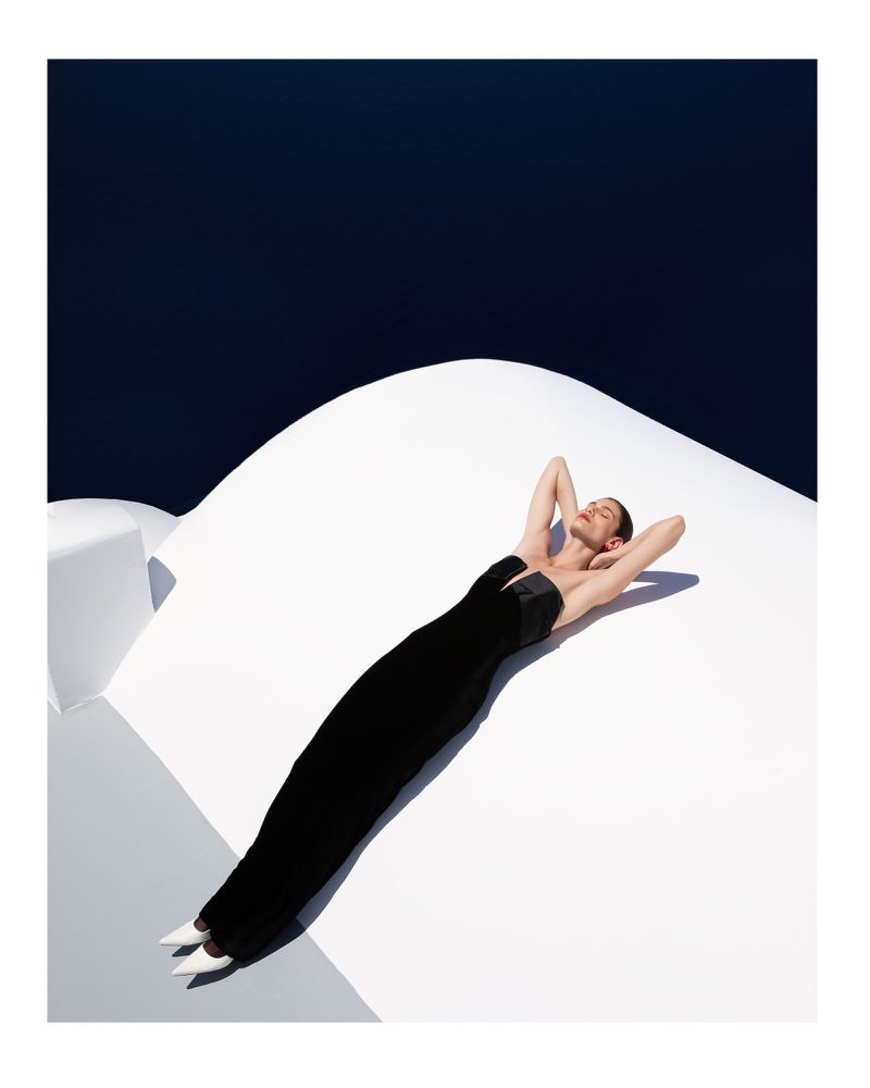 Woman in black dress lying on white rooftop against dark blue sky. Fashion shot with contrasting colors and modern minimalist style.