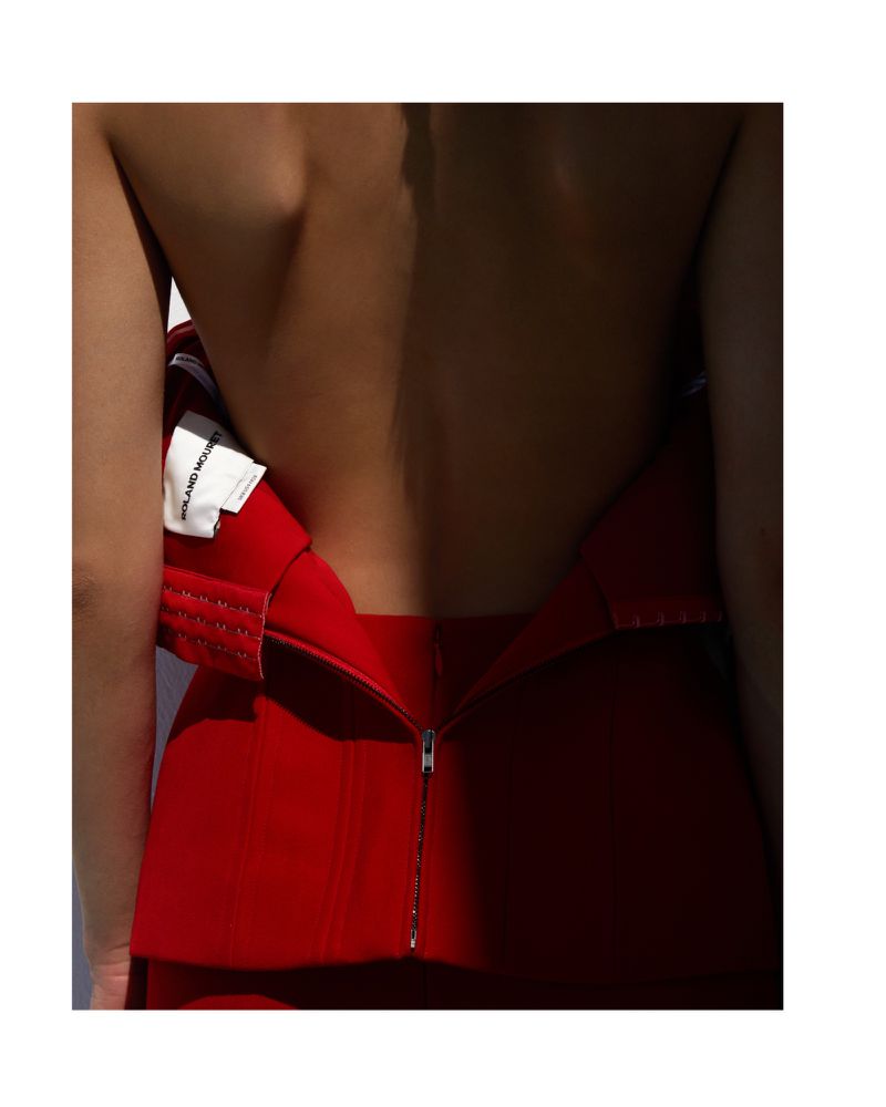 Close-up of woman's back and red dress. The dress has a zipper. Part of a white label is visible. Soft light casts shadows across the body.