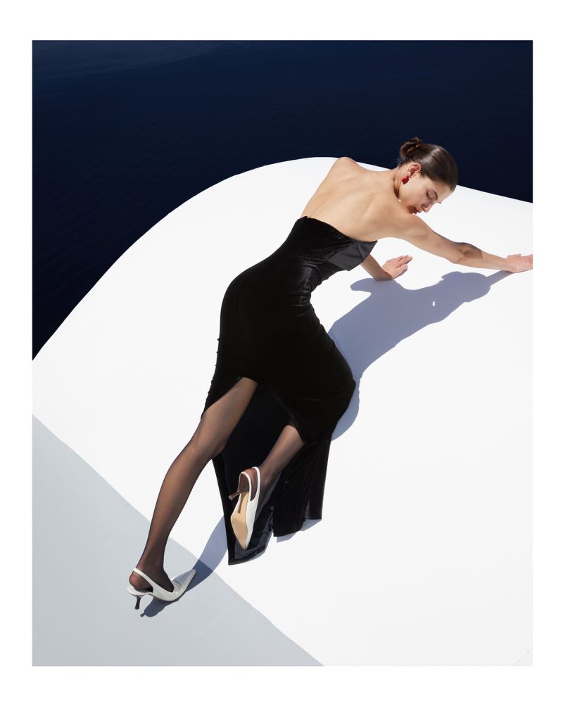 Fashion editorial shot of a woman in a black gown and sheer hose, posed on a white surface, with a dark blue background.