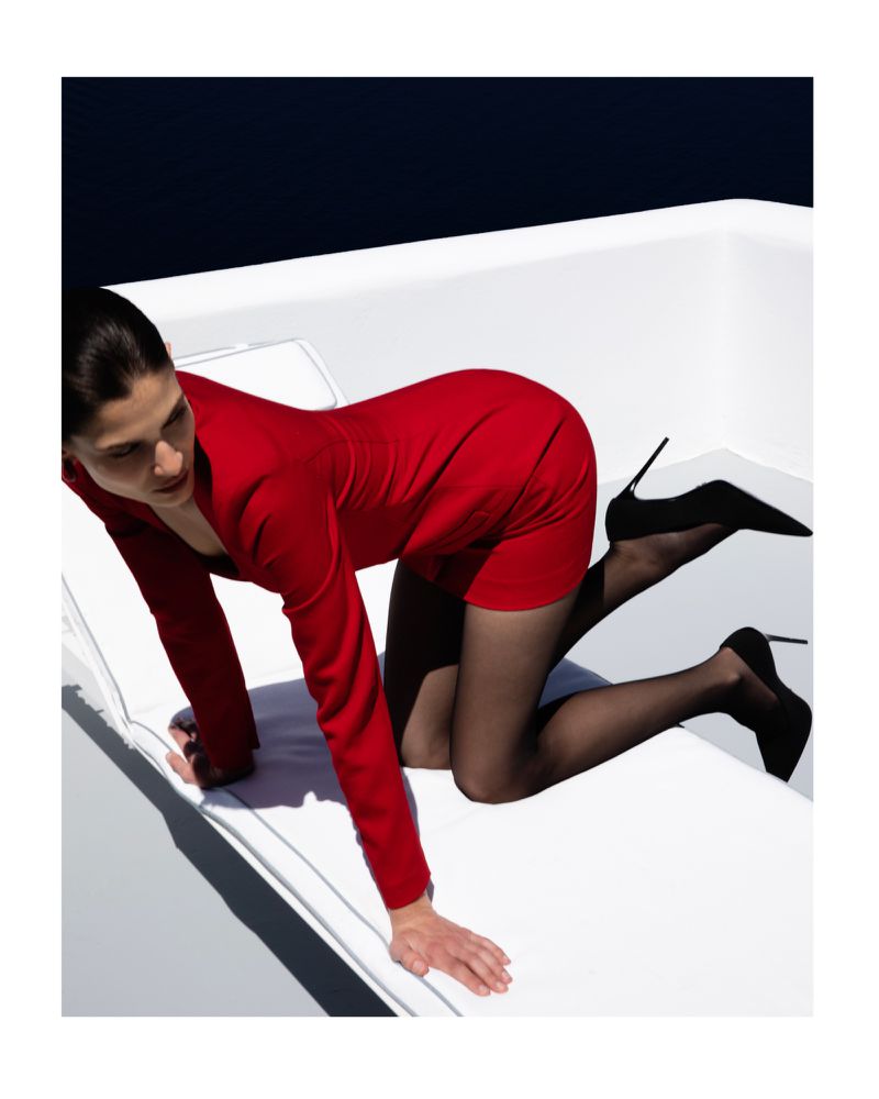Woman in red dress and black heels posing on white surface against dark blue background. Fashion, portrait, studio shot.