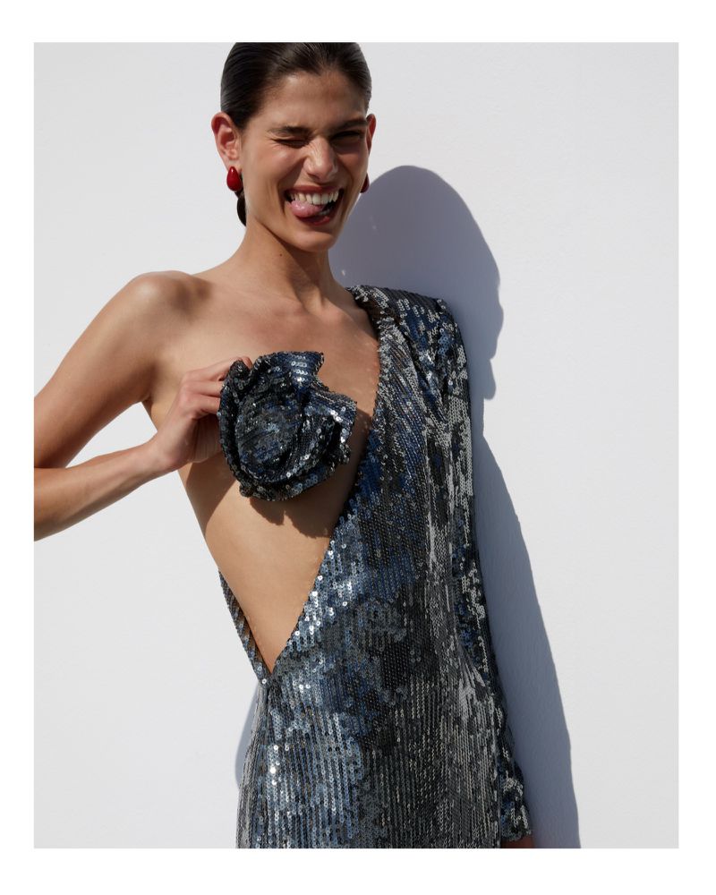 Brunette woman in a blue sequin one-shoulder dress posing against a white wall with tongue out, accessorized with red earrings.