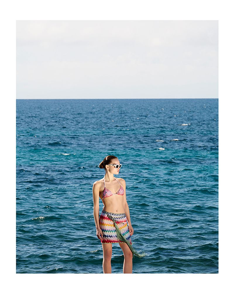 Woman in bikini and skirt stands against the ocean backdrop. Beach fashion, swimwear, and summer style. Coastal lifestyle.