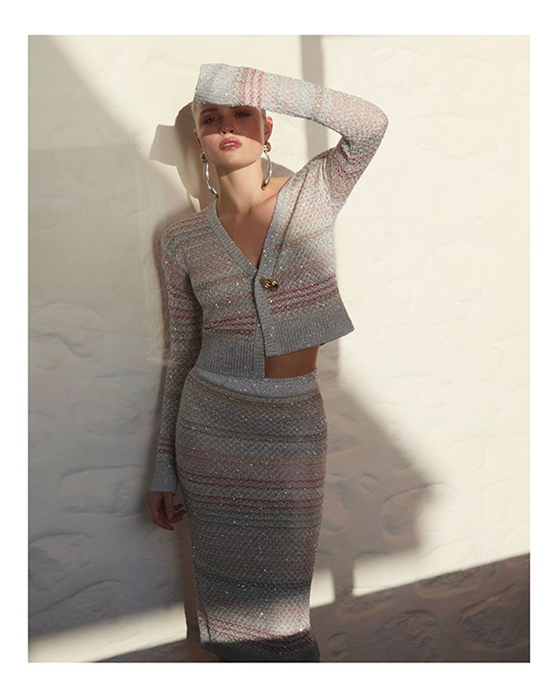 Model leaning against a textured stone wall wearing a patterned knit cardigan and matching midi skirt.