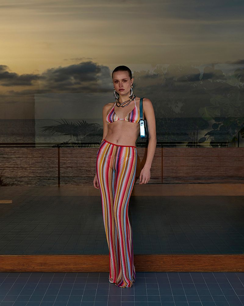 Fashionable adult woman poses in a multicolored knit bikini and pants with a modern handbag against a sunset ocean view.